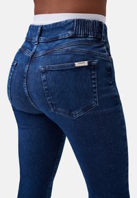 Jeans in denim blu scuro con vita alta, dotati di un pannello elastico arricciato, tasche posteriori a toppa e dettagli di cucitura minimi.