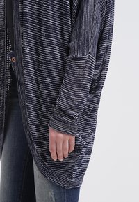 Cardigan a righe navy con vestibilità ampia, maniche lunghe e fondo arrotondato. Presenta un motivo testurizzato e un bottone sul davanti.