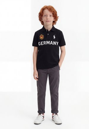 Rudowłosy chłopiec ubrany w czarne polo z napisem „GERMANY”, szare spodnie, białe tenisówki, stojący z jedną ręką w kieszeni na białym tle.