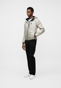 Άνδρας που φοράει ανοιχτό γκρι μπουφάν με κουκούλα, μαύρο παντελόνι και λευκά αθλητικά παπούτσια, στέκεται με το ένα χέρι στην τσέπη.