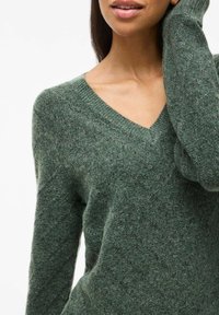 Pull vert en col V, fabriqué en tissu tricoté texturé, avec des poignets côtelés et une coupe décontractée. Motif diagonal subtil sur la surface.