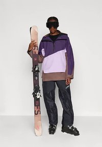 Osoba ubrana w fioletowy, brązowy i czarny strój narciarski z czarnymi butami narciarskimi i goglami, trzymająca pionowo parę różowo-czarnych nart.