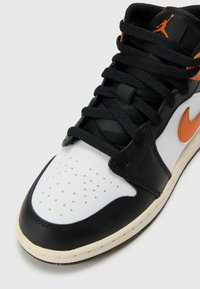 Svart og hvit lærsneaker med oransje detaljer, utstyrt med perforert tå, runde snøring og en strukturert overflate på overdelen.