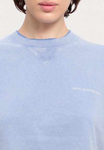 Light blue sweatshirt with a crew neckline, featuring raw edges. Subtle white branding on the chest. Soft fabric with a slightly textured finish.