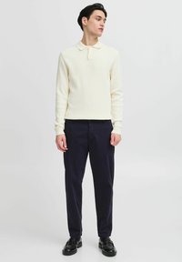 Cream ribbed long-sleeve polo shirt with a collar, paired with navy trousers and black shoes, standing against a plain background.