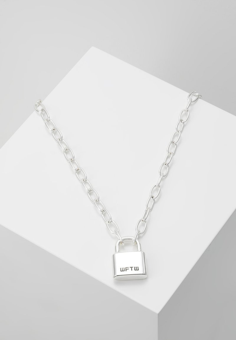 Collier chaîne en argent avec un pendentif cadenas rectangulaire gravé de "WFTW" sur une surface d'exposition blanche inclinée.