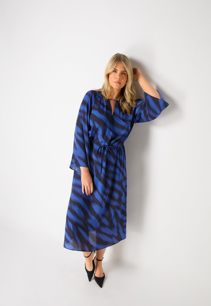 Modré a černé šaty se vzorem, s úzkým pasem, dlouhými rukávy a výstřihem do V. Vyrobené z lehkého, splývavého materiálu.