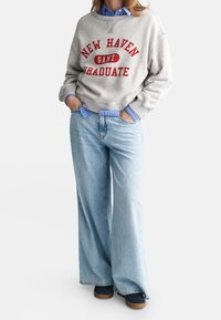 Sudadera gris con texto "NEW HAVEN GRADUATE" en rojo. Combinada con vaqueros de pierna ancha en azul claro. Con una camisa a rayas azules debajo.