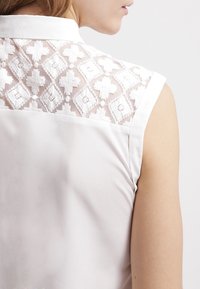 Blusa bianca con un pannello in pizzo testurizzato con motivi geometrici sul retro. Maniche a cappuccio e un tessuto leggero e morbido.