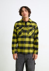 Grøn og sort ternet flannel skjorte med knaplukning fortil, to brystlommer og lange ærmer, parret med grå bukser.