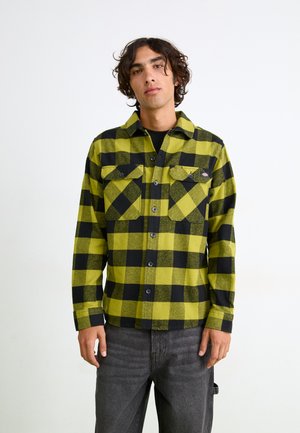 Green and black checkered flannel shirt with button-down front, two chest pockets, and long sleeves, paired with gray pants.