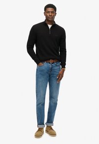 Suéter negro con cremallera, jeans azules, cinturón marrón y zapatos beige con puños enrollados. Textura suave y diseño ajustado. Look sencillo y clásico.