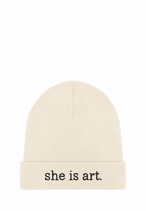 Bonnet tricoté en beige clair avec un revers. Présente un texte brodé en noir indiquant "she is art." La texture est douce et extensible.