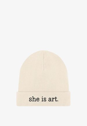 Bonnet tricoté en beige clair avec un revers. Présente un texte brodé en noir indiquant "she is art." La texture est douce et extensible.