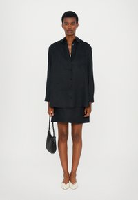Black oversized shirt with button-down front and pocket, paired with a matching skirt. Model holds a textured black handbag and wears cream pointed shoes.
