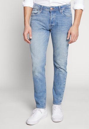 Mand iført lyseblå slim-fit jeans og hvide sneakers, stående med hænder afslappet ned langs siderne mod en ensfarvet baggrund.