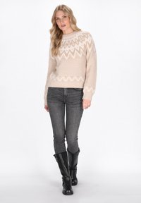 Ragazza con lunghi capelli biondi che indossa un maglione beige a fantasia, jeans skinny grigi e stivali neri fino al ginocchio, in piedi contro uno sfondo bianco.