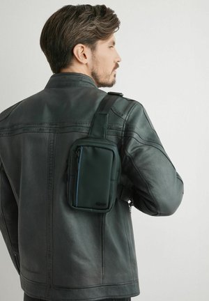 Mężczyzna ubrany w czarną skórzaną kurtkę, z małą czarną torebką na ramię przewieszoną przez prawy bark, zwrócony w prawo na jednolitym tle.