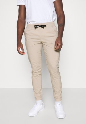 Mand iført beige slim-fit løbebukser med sort snøre, hvide sokker, hvide sneakers og en hvid t-shirt mod en ensfarvet lys baggrund.