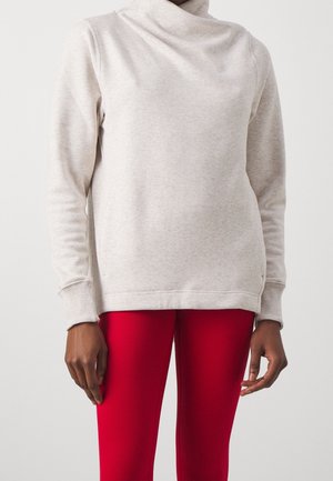 Personne portant un sweat-shirt beige clair à manches longues avec un col montant et un pantalon côtelé rouge vif sur un fond uni.