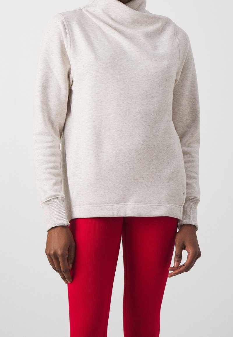 Personne portant un sweat-shirt beige clair à manches longues avec un col montant et un pantalon côtelé rouge vif sur un fond uni.
