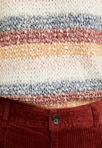 Jersey de punto con rayas multicolores en blanco, azul, rojo y amarillo, combinado con pantalones de pana burdeos texturizados que cuentan con dos botones negros.