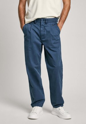 Blue cotton trousers with a relaxed fit, featuring front pleats, side pockets, and a button closure. Paired with white sneakers.