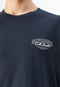 Mörkblå crewneck t-shirt med vit text "JACK & JONES Coll. Studio Created by the J&J crew Since 1990" på vänster bröst.