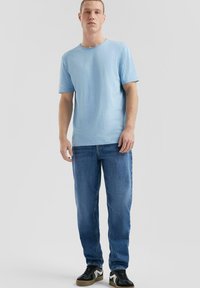 Jasnoniebieska bawełniana koszulka z krótkimi rękawami, okrągłym dekoltem, noszona z niebieskimi jeansami i czarnymi trampkami z białymi i szarymi akcentami.