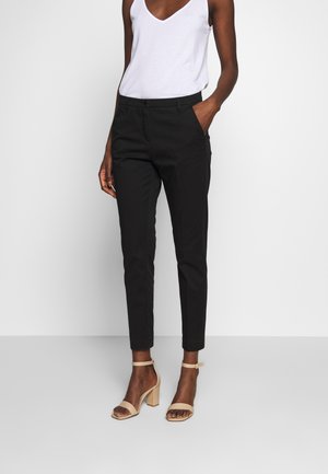 Vrouw draagt zwarte, nauwsluitende enkellange broek, wit mouwloos topje en beige sandalen met hoge hak, staand tegen een effen achtergrond.