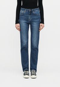 Άτομο που φοράει σκούρα μπλε τζιν με ίσια γραμμή, μαύρο μακρυμάνικο μπλουζάκι και μαύρα-λευκά αθλητικά παπούτσια, στέκεται μπροστά σε απλό λευκό φόντο.