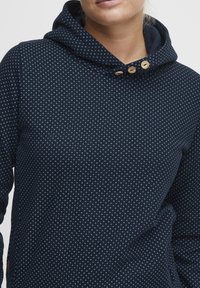 Hoodie bleu marine avec un motif moucheté, fabriqué en matériau doux. Doté d'une capuche avec cordon de serrage et d'accents en bois près de l'encolure.