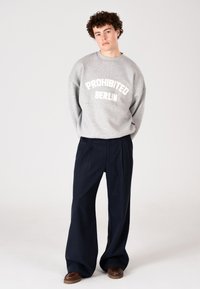 Fiatal férfi göndör hajjal, szürke "PROHIBITED BERLIN" pulóverben, széles fekete nadrágban és barna cipőben, egy sima háttér előtt áll.