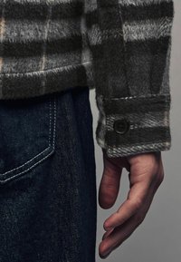 Camisa de franela gris a cuadros con un botón negro en el puño, combinada con unos vaqueros de mezclilla azul oscuro que cuentan con un bolsillo frontal y detalles en costura blanca.