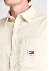 Beige kordflämskjorta med en button-down krage, med en bröstficka och ett märke med en röd, vit och blå logotyp. Texturerad tyg.