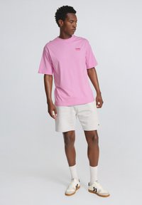 Lyserød bomulds t-shirt med et lille rødt logo, parret med lysegrå shorts og hvide sneakers med sorte detaljer og sokker.