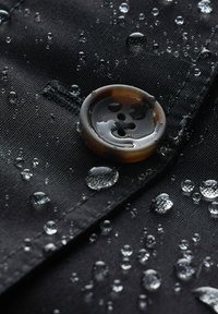 Tessuto nero con gocce d'acqua; presenta un bottone marrone lucido con quattro fori, superficie texturizzata e cuciture visibili.