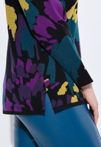 Nahaufnahme eines bunten, floral gemusterten Pullovers mit lila, gelben und türkisfarbenen Formen, der über blauen Lederhosen getragen wird.