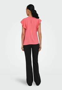 Blusa rosa corallo con maniche a volant e dettaglio a chiave sul retro, abbinata a pantaloni neri a gamba larga. Il tessuto sembra leggero con una finitura testurizzata.