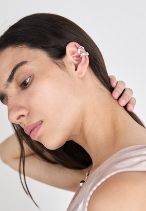 Stříbrný ušní šperk s několika ryzími kamennými akcenty, nošený na horní části ucha. Hladký povrch, moderní design, částečně zakrytý dlouhými vlasy.