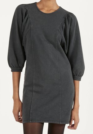 Robe gris foncé ajustée avec manches trois-quarts bouffantes et coutures verticales visibles sur le devant, portée avec des collants noirs.
