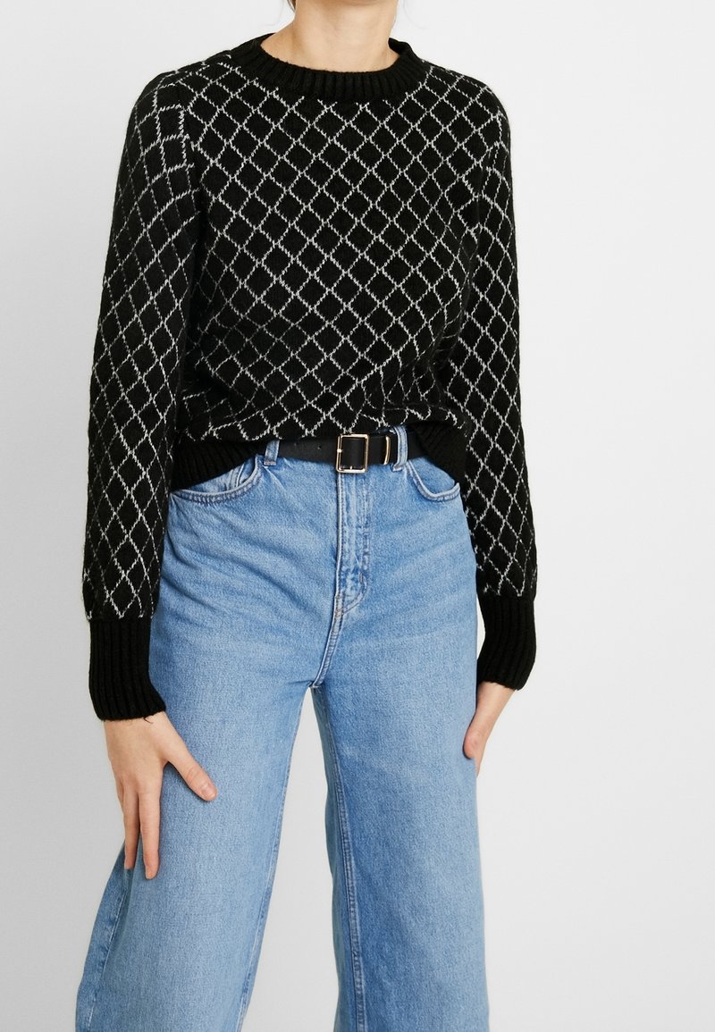 Personne portant un pull noir avec un motif losange blanc et un jean bleu clair taille haute avec une ceinture noire, debout les mains détendues.