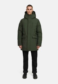 Donkergroen gewatteerde parka met capuchon, ritssluiting, twee voorzakken en een gestructureerde afwerking. Draagt over zwarte broek en laarzen.