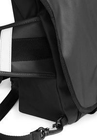 Svart budväska med en texturerad tyg, justerbar rem och kontrasterande vit rand. Robust rektangulär form med detaljerad sömnad.