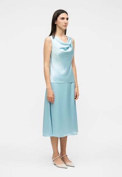 Γυναίκα που φοράει μπλε-ανοιχτό σατέν αμάνικο τοπ και αντίστοιχη μίντι φούστα, με μπεζ μυτερές φλατ παπούτσια, σε απλό φωτεινό φόντο.