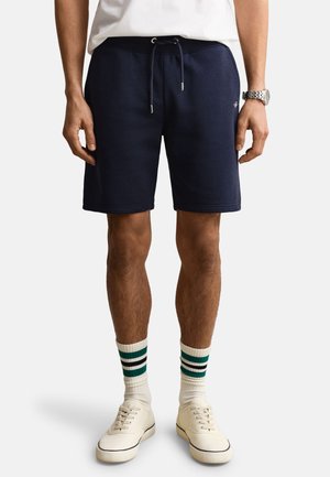 Mann iført marineblå shorts med snor, hvite stripete sokker, hvite joggesko, en hvit skjorte og et sølvfarget armbåndsur, stående på hvit bakgrunn.