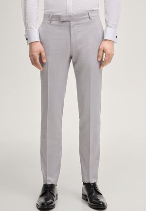 Uomo che indossa pantaloni sartoriali grigio chiaro, camicia bianca elegante e scarpe nere lucide, in piedi contro uno sfondo semplice.