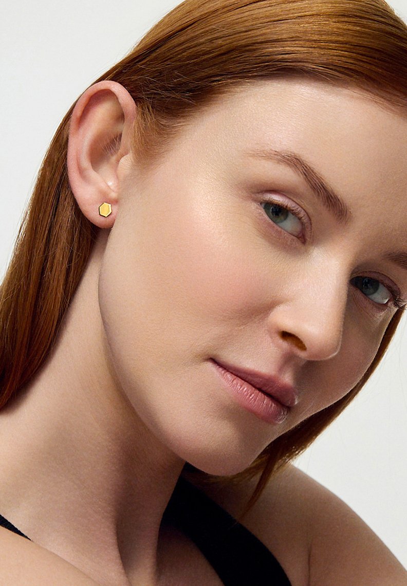Złoty sześciokątny kolczyk wkrętka, mały rozmiar, gładka powierzchnia, minimalistyczny design, na neutralnym tle, noszony na uchu modelki.