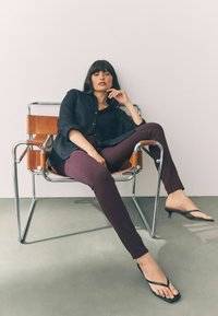 Zwarte button-up shirt, bordeaux fitted broek en zwarte heeled flip-flops. Model zit in een chromen stoel met leren accenten tegen een lichte muur.