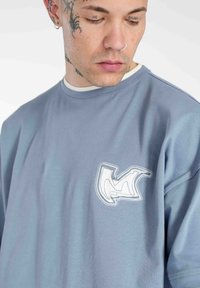 Blå bomulds sweatshirt med rund hals, der har et hvidt broderet logo på brystet. Stoffet har en glat tekstur og en afslappet pasform.